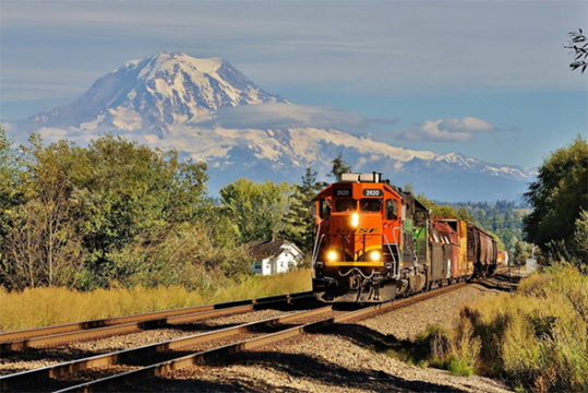 The FRA continues to focus on Positive Train Control (PTC) implementation with a new push to speed progress as well as the release of more grant funding. A BNSF train speeds along main line track that will be part of the PTC network near Puyallup, Washington beneath Mount Rainier. Image © 8/2016 by Nikki Burgess; all rights reserved.